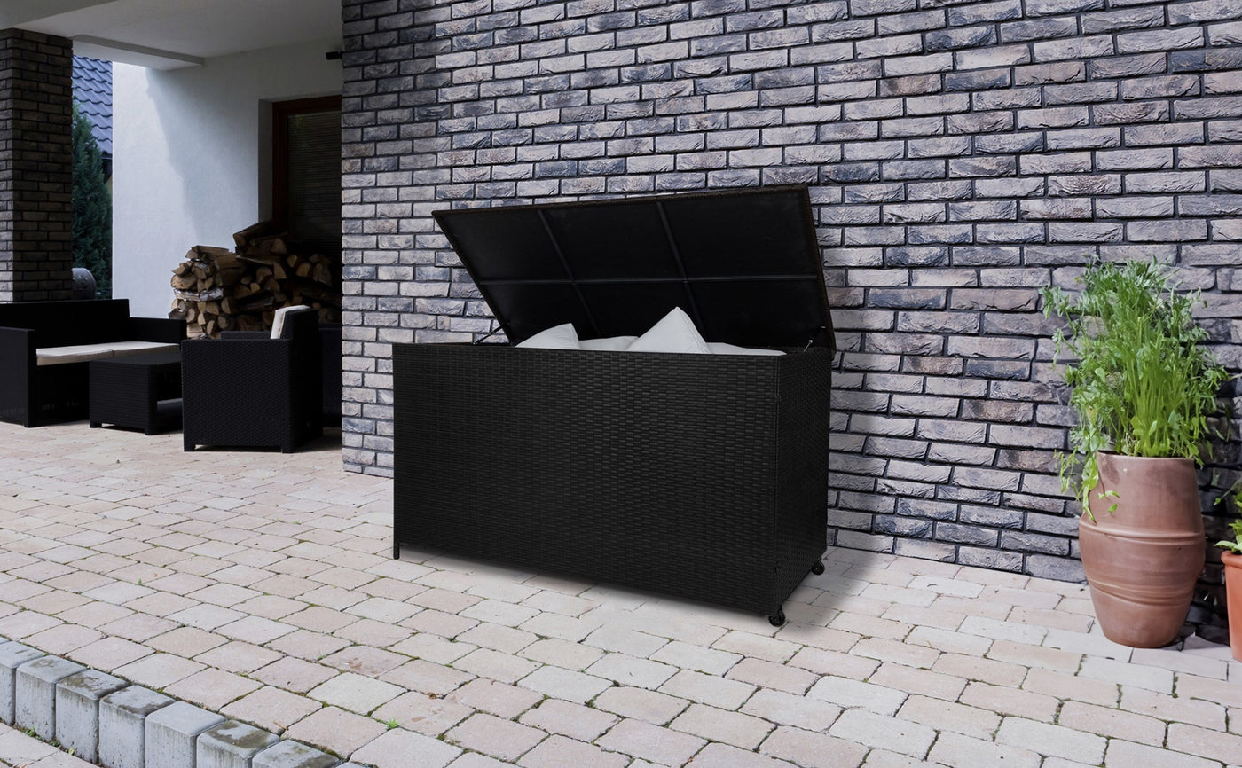 Coffre de rangement noir en résine tressée, ouvert, avec coussins blancs à l'intérieur, sur un patio en pavés, mur en briques en arrière-plan