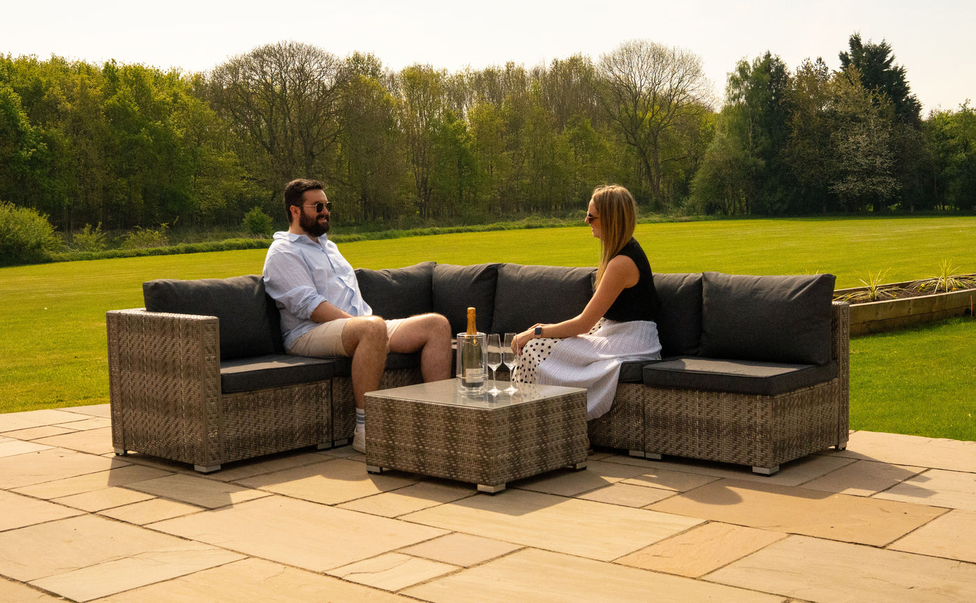 Canapé en rotin tissé avec coussins gris, deux personnes assises, un verre d'eau et une bouteille de champagne sur une table basse, jardin verdoyant en arrière-plan