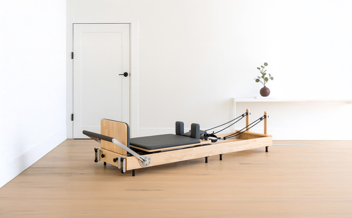 Reformer de Pilates en bois clair avec un coussin noir, cordes élastiques et roulettes, dans une pièce minimaliste lumineuse