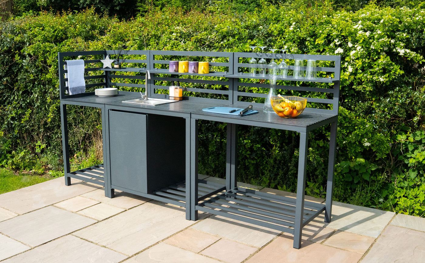 Bar de jardin moderne en métal noir avec plan de travail, évier, et décorations colorées, entouré de verdure et de pavés clairs