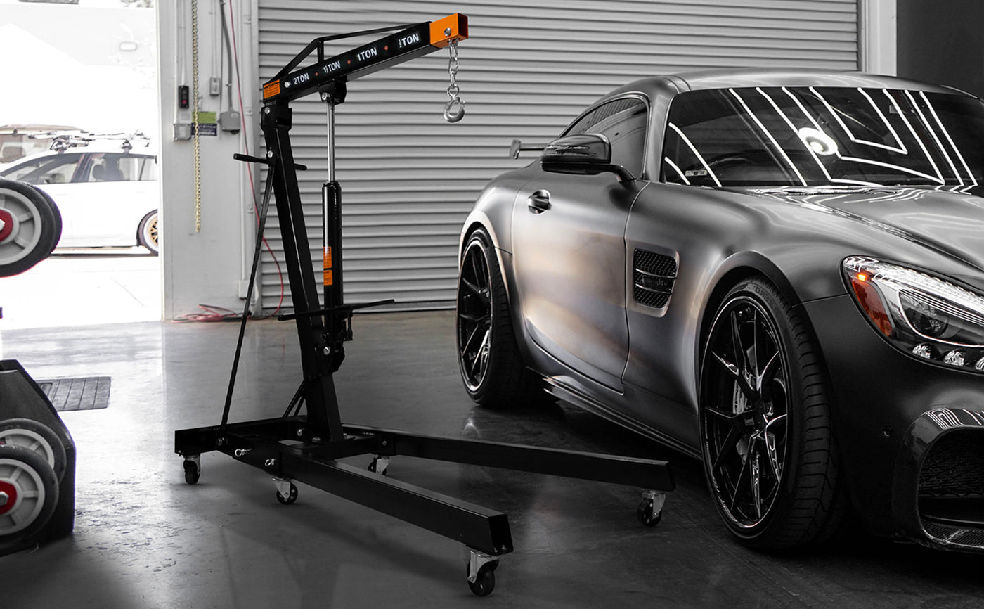 Un palan noir et orange à roulettes à côté d'une voiture sportive matte grise, avec des reflets brillants et des jantes noires élégantes