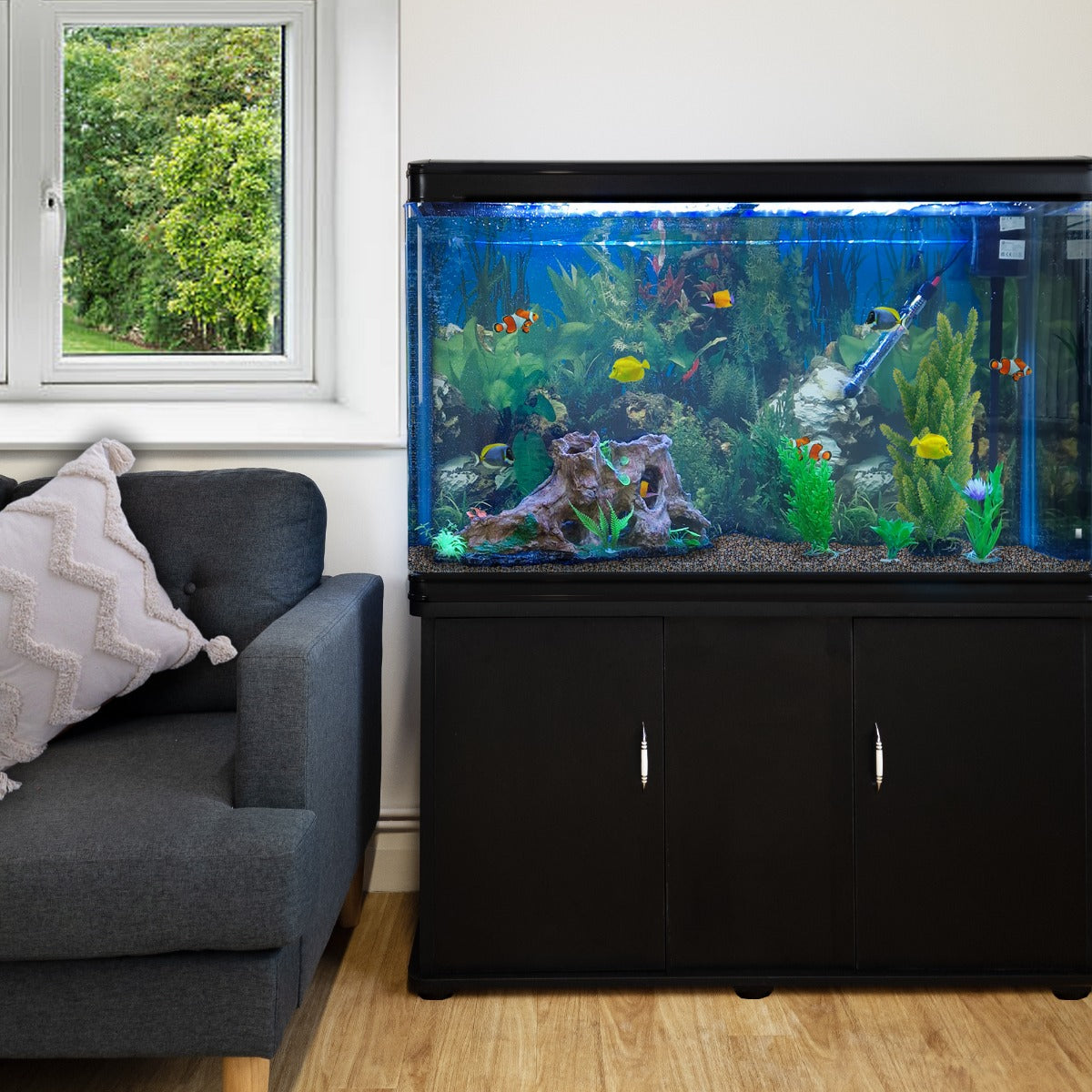 Aquarium noir avec meuble de support, gravier naturel, plantes aquatiques colorées et poissons, à côté d'un canapé gris et d'une fenêtre