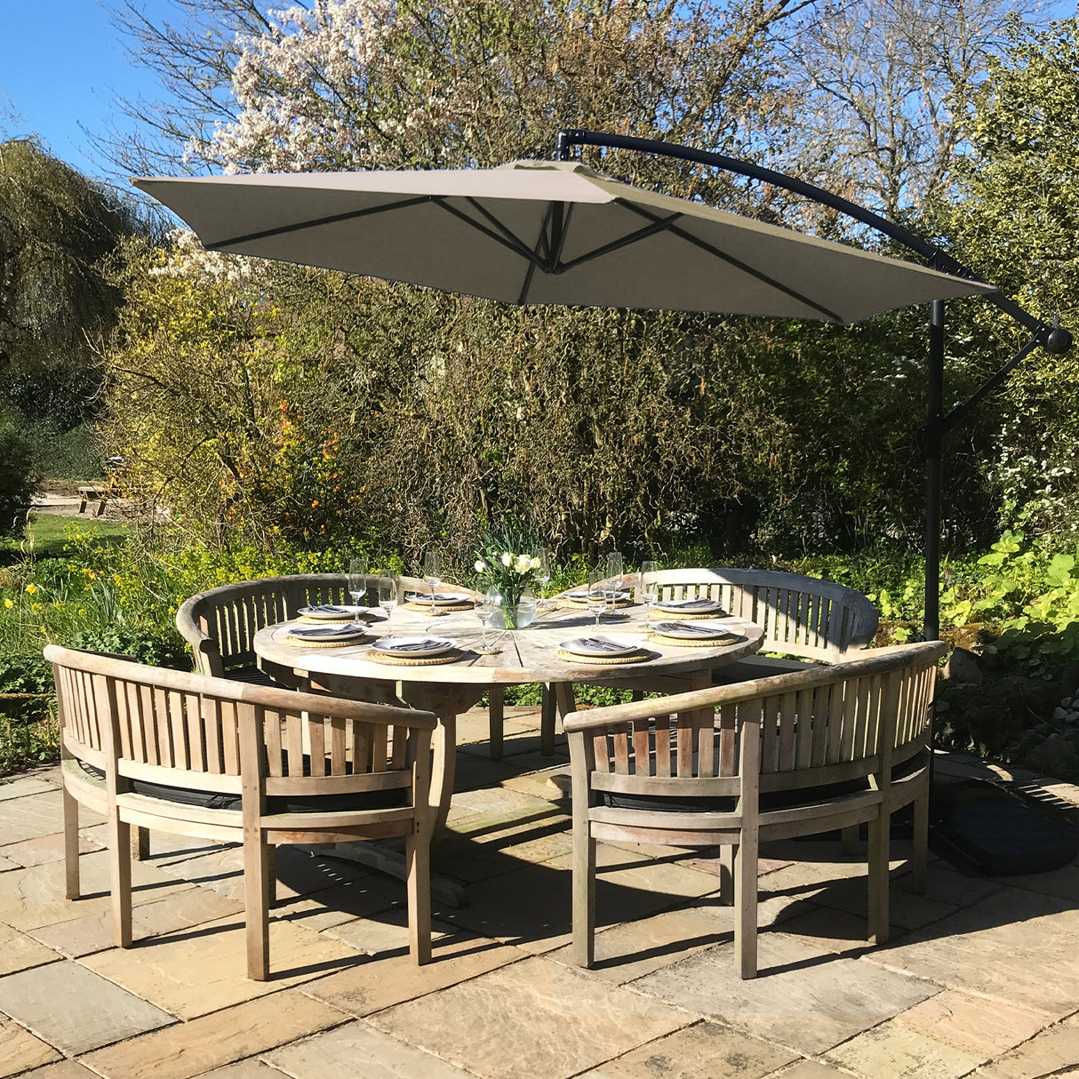 Parasol déporté beige de 3 m au-dessus d'une table ronde en bois entourée de chaises, dans un jardin verdoyant ensoleillé