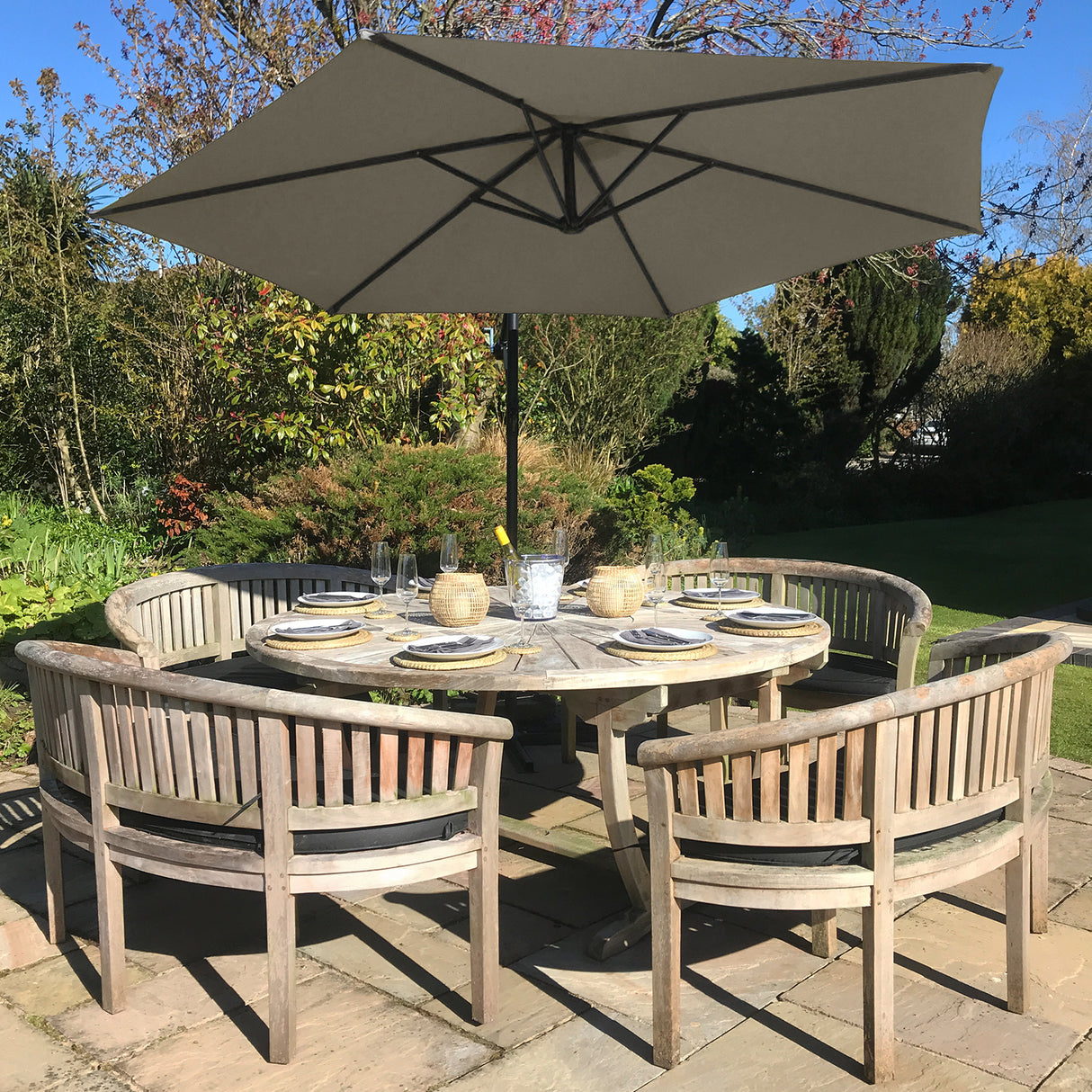 Parasol déporté beige de 3 m sur une table en bois entourée de chaises, dans un jardin verdoyant avec des fleurs en arrière-plan
