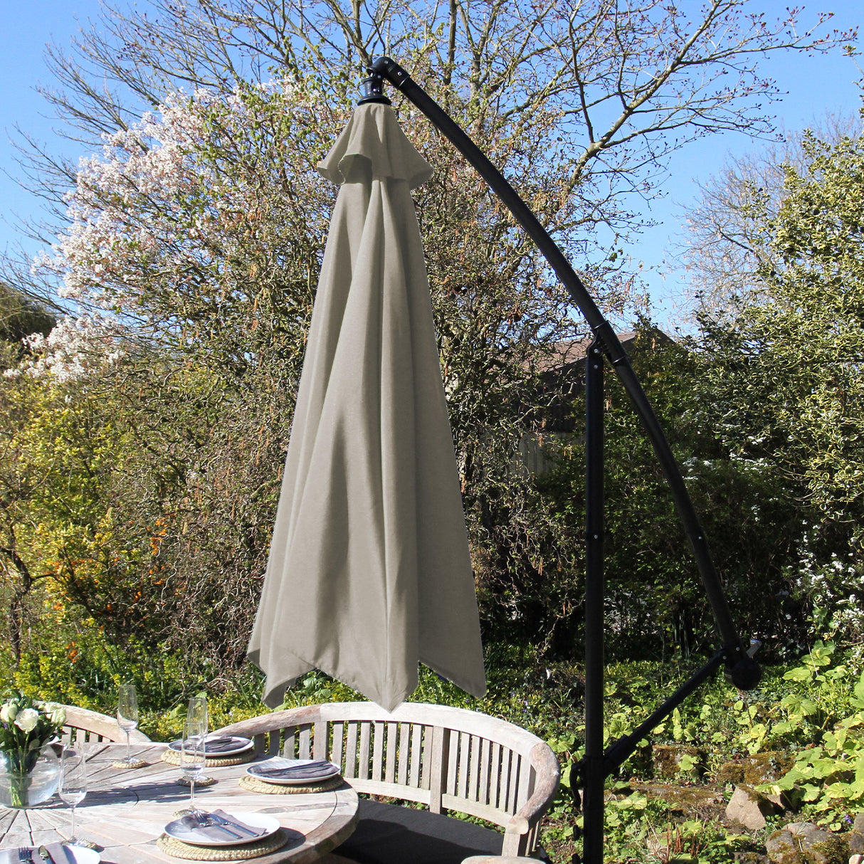 Parasol déporté de 3 m en beige, avec une structure noire élégante, installé au-dessus d'une table en bois dans un jardin verdoyant