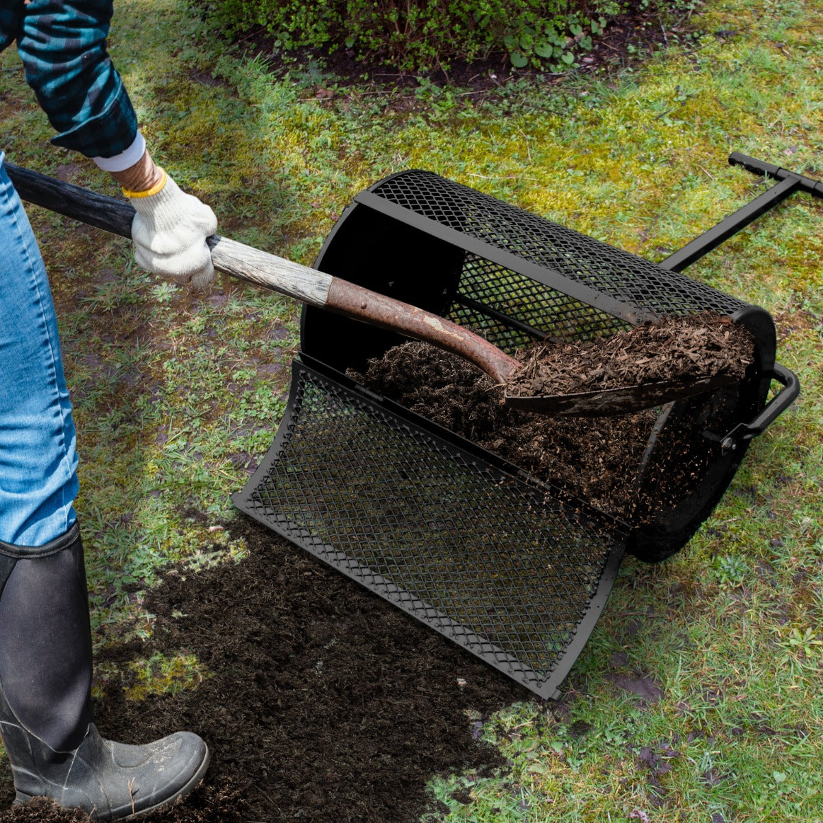 Rouleau épandeur à compost noir de 80 L, avec texture en métal perforé, déversant du compost sur une pelouse verte et humide