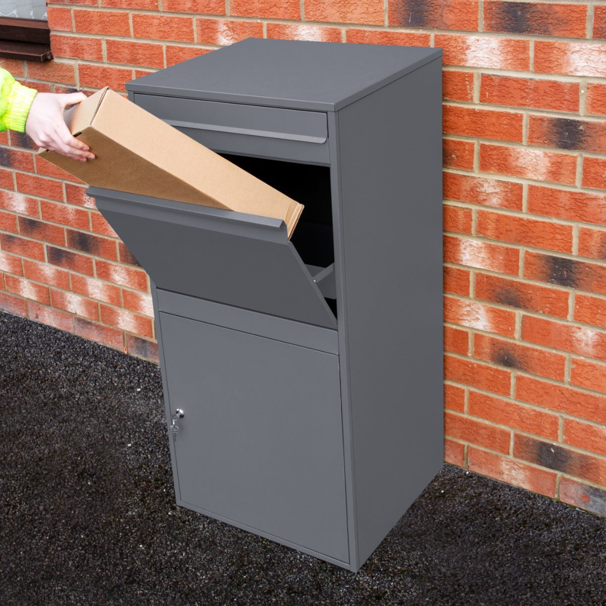 Boîte à colis XL grise, en métal, avec une ouverture supérieure pour les colis et un compartiment verrouillable en bas, placée contre un mur en briques