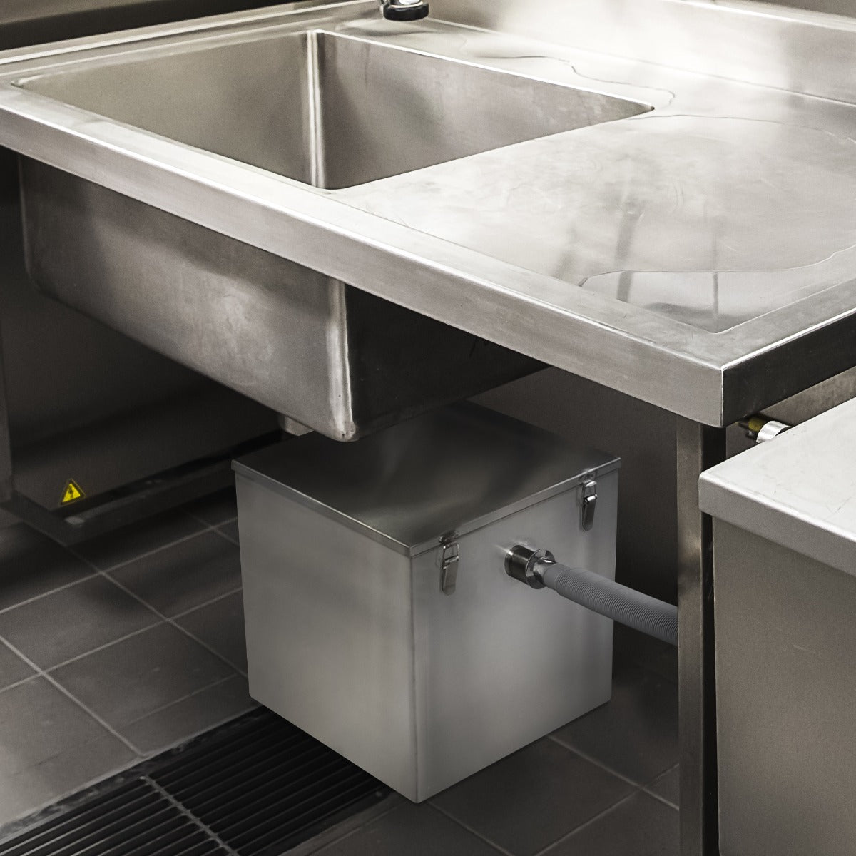 Bac à graisse commercial KuKoo de 60L en acier inoxydable, positionné sous un évier de cuisine, avec un tuyau de drainage connecté