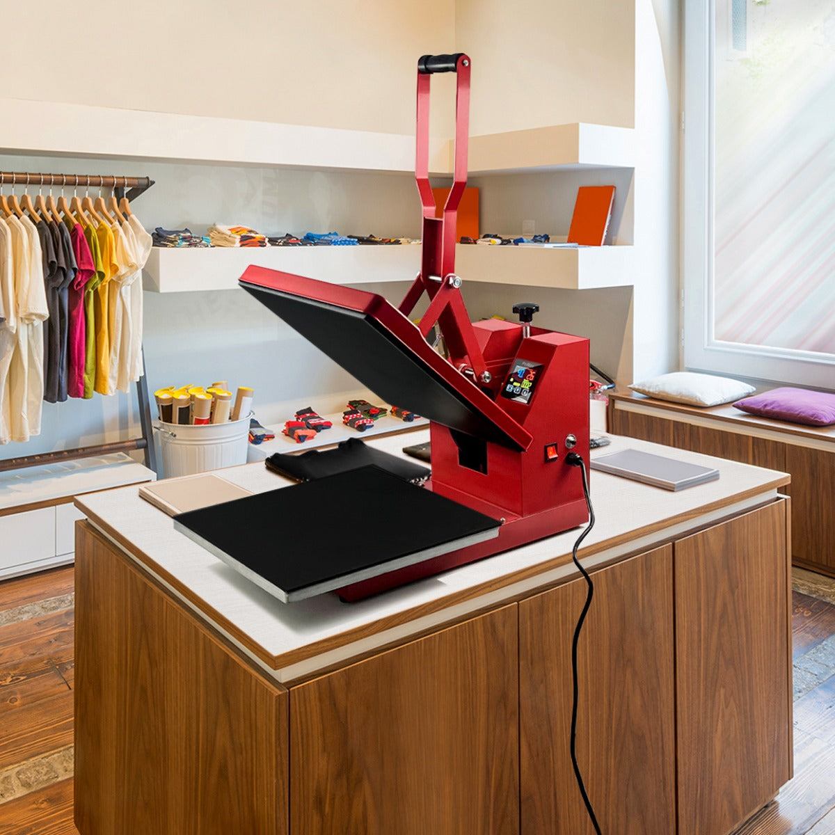 Presse à chaud rouge de 38 cm avec plateau noir, placée sur un meuble en bois, entourée de vêtements colorés et d'accessoires