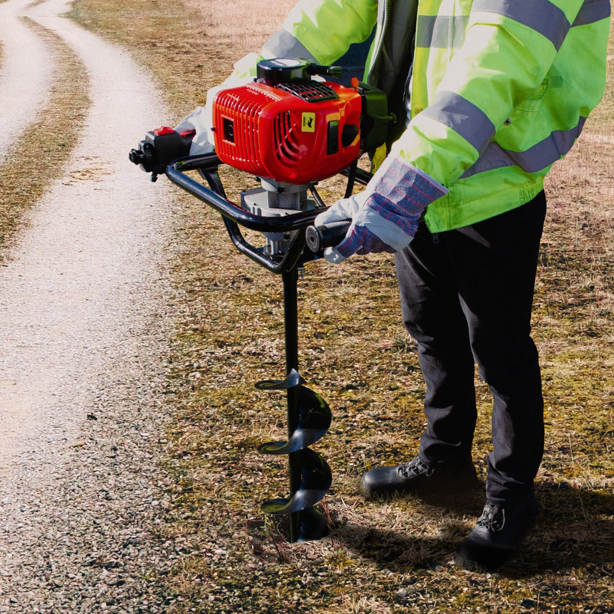 Tarière T-Mech 52cc 5 en 1, avec moteur rouge, utilisée sur un sol herbeux, homme en veste réfléchissante et gants, en action