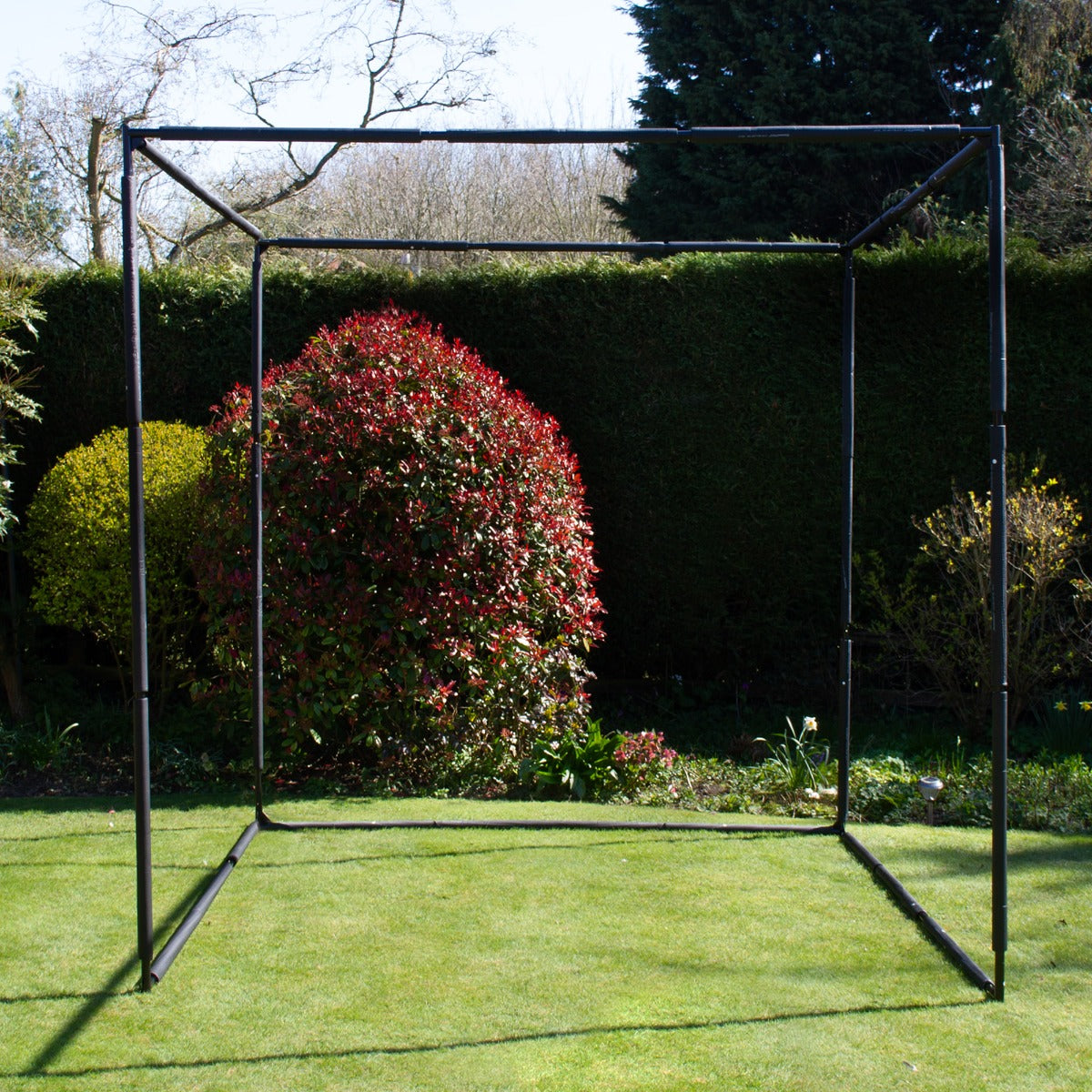 Cage d'entraînement de golf en métal noir, structure carrée, installée sur une pelouse verte, entourée de buissons fleuris rouges et verts