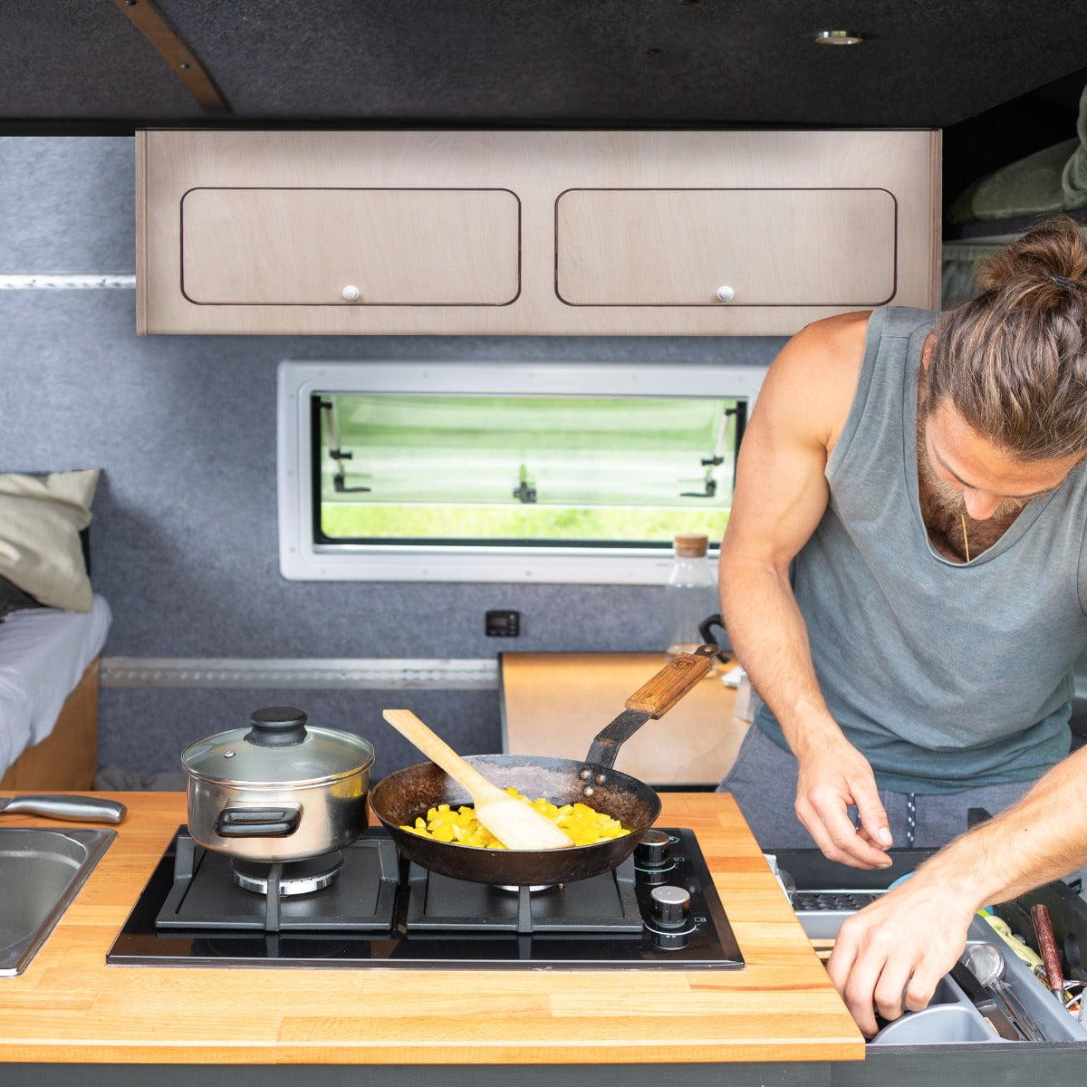 Un homme cuisine dans un camping-car, avec une armoire de rangement en bois clair, un plan de travail en bois et des ustensiles de cuisine en acier inoxydable