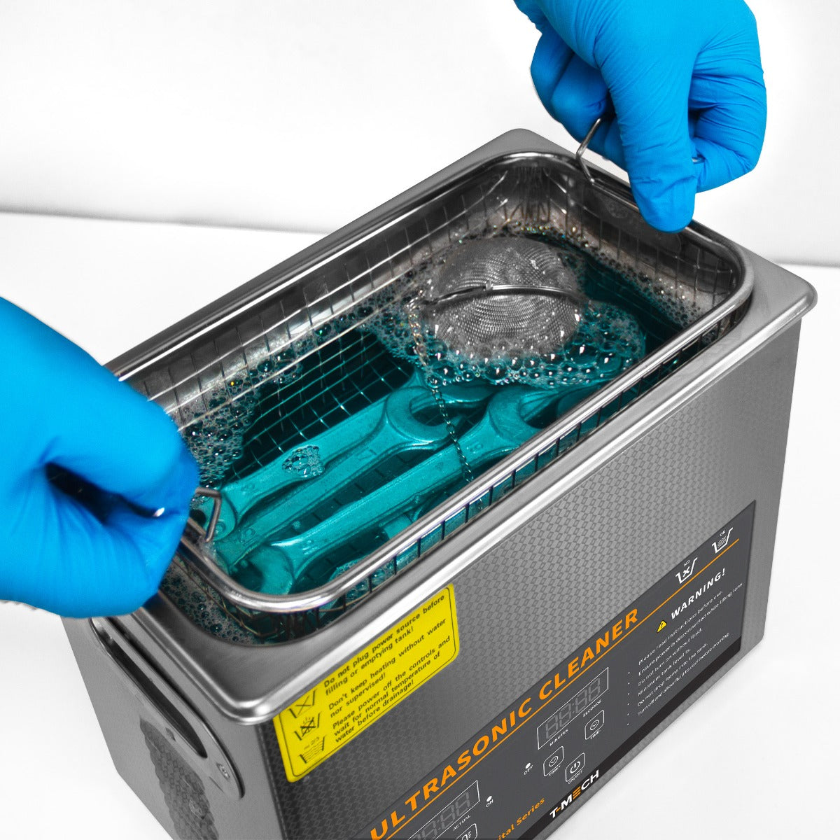 Nettoyeur ultrasonique T-Mech de 3 L avec un panier en métal, eau bleue pétillante et mains en gants bleus manipulant l'équipement