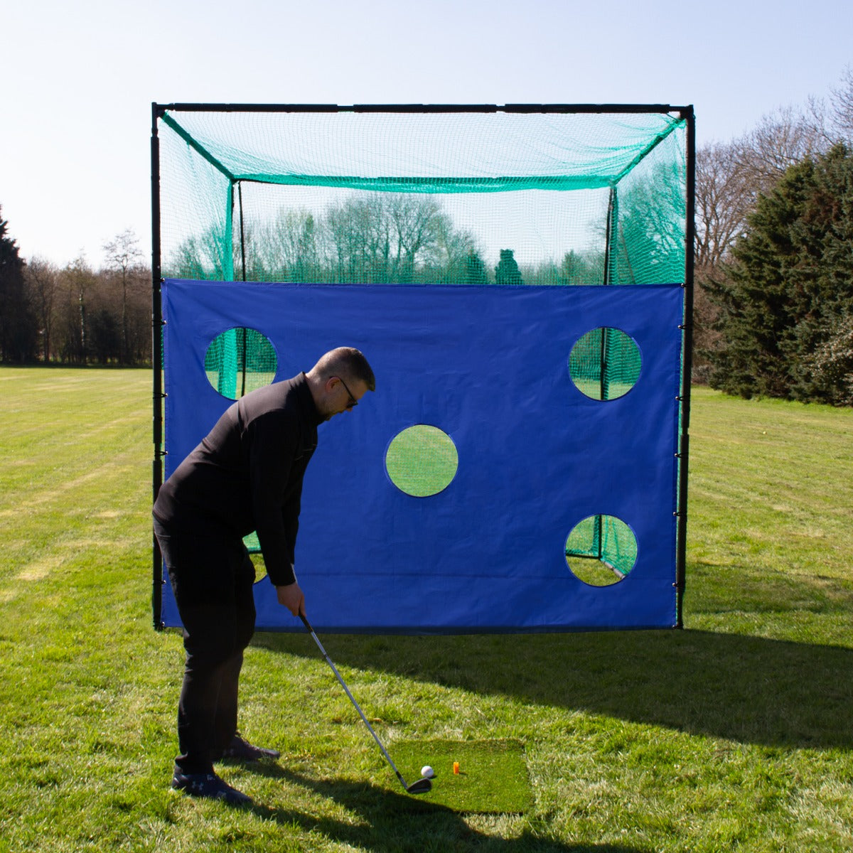 Cible multi-trous bleue pour l'entraînement de golf, avec filet vert, installée sur un terrain ensoleillé, joueur prêt à frapper la balle