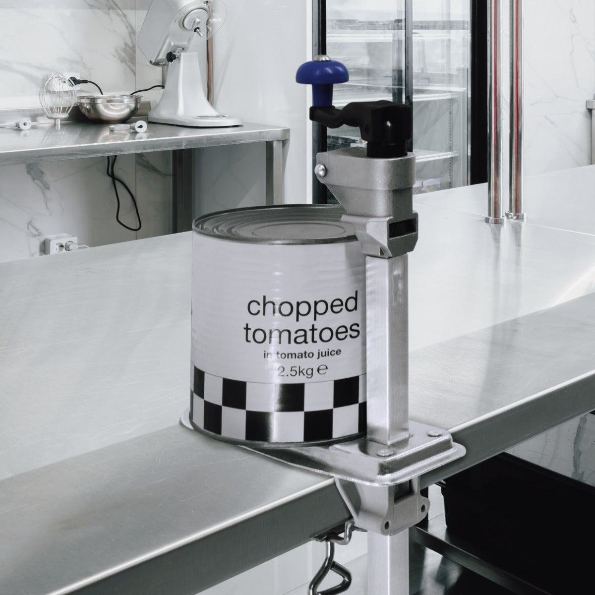 Ouvre-boîte commercial en métal argenté avec un bouton bleu, tenant une boîte de tomates hachées sur une surface de travail en acier inoxydable