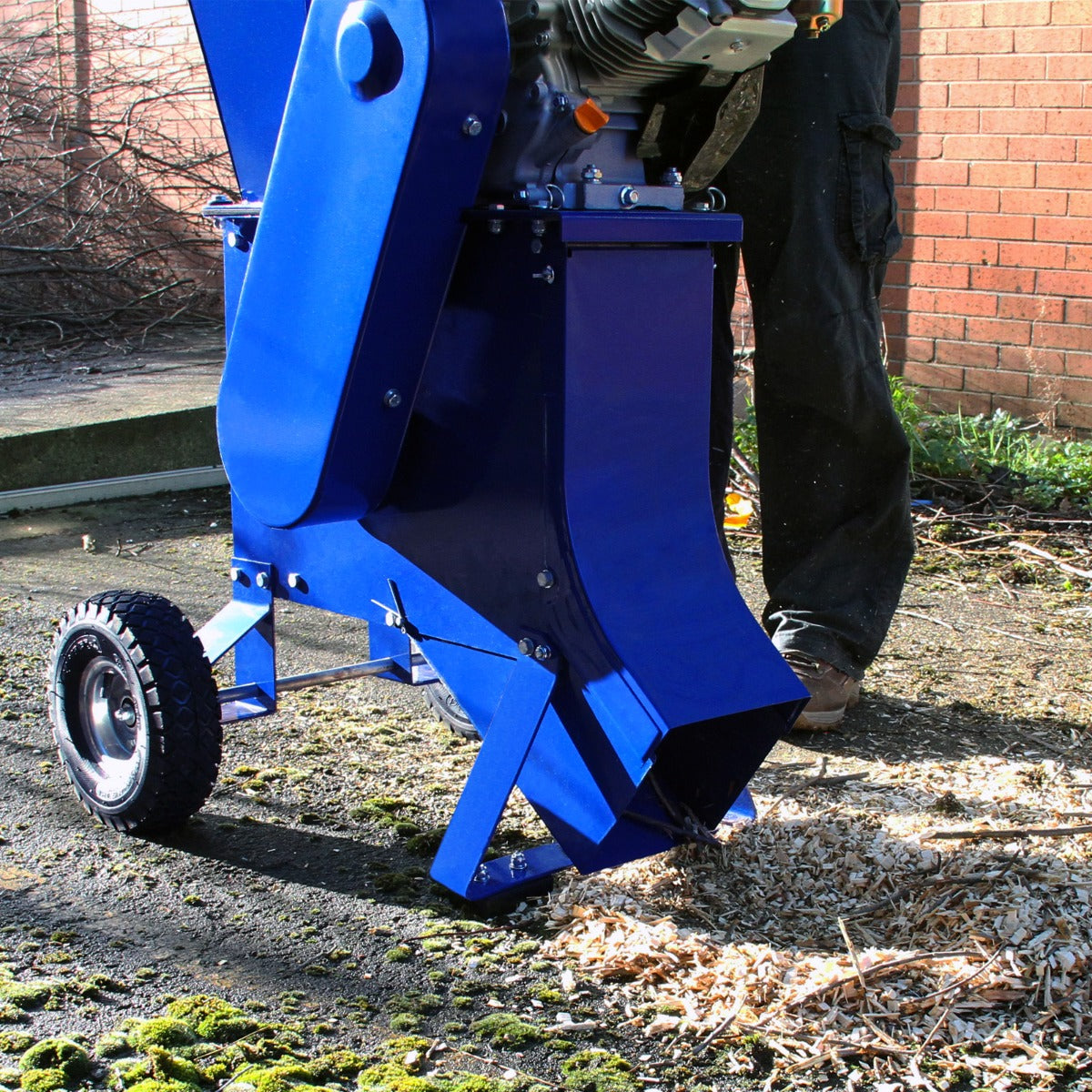 Déchiqueteuse de bois 7CV en métal bleu, avec roues, broyant des copeaux de bois sur un sol en béton, en arrière-plan de briques rouges