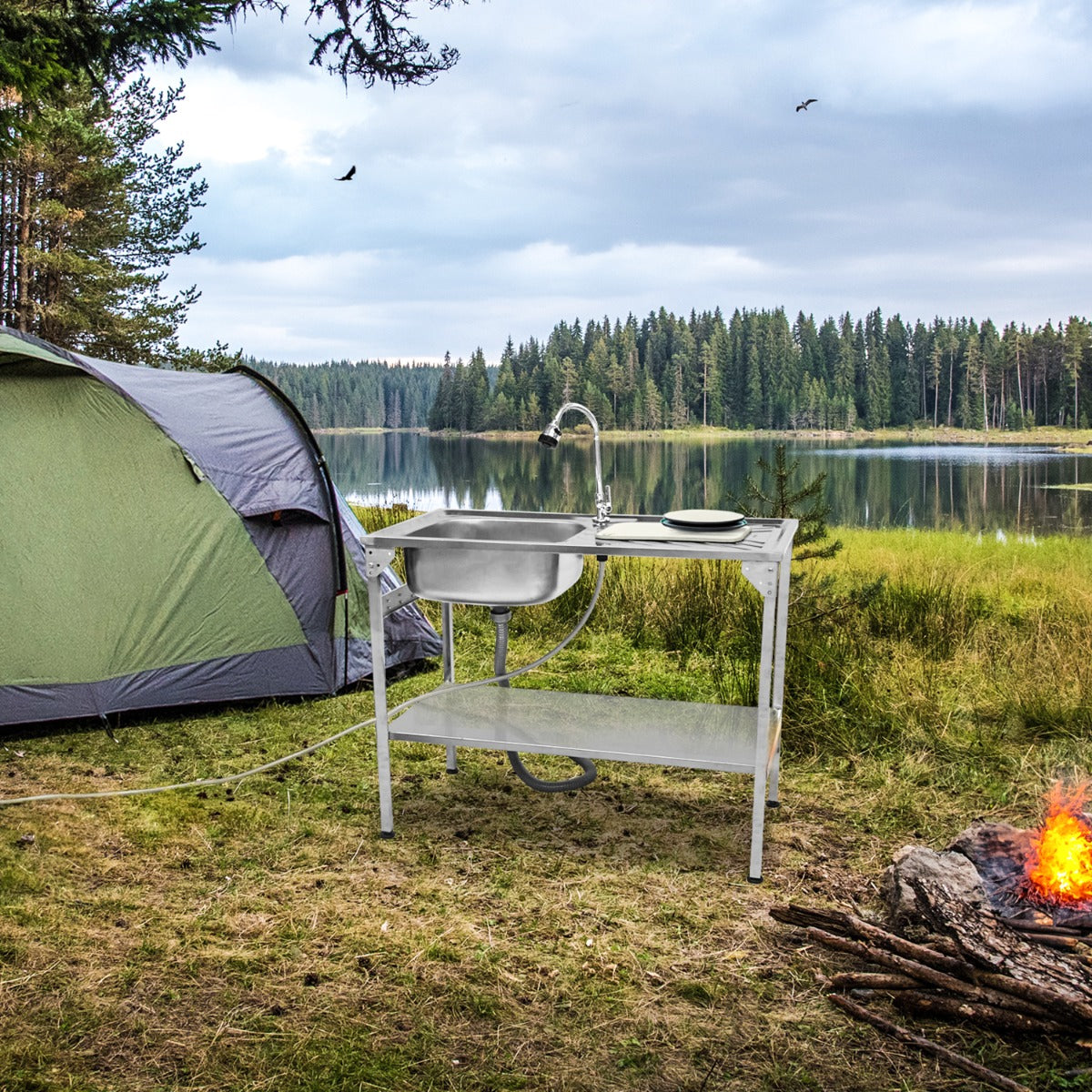Évier de camping pliable en métal argenté, avec robinet, installé près d'une tente verte au bord d'un lac entouré d'arbres