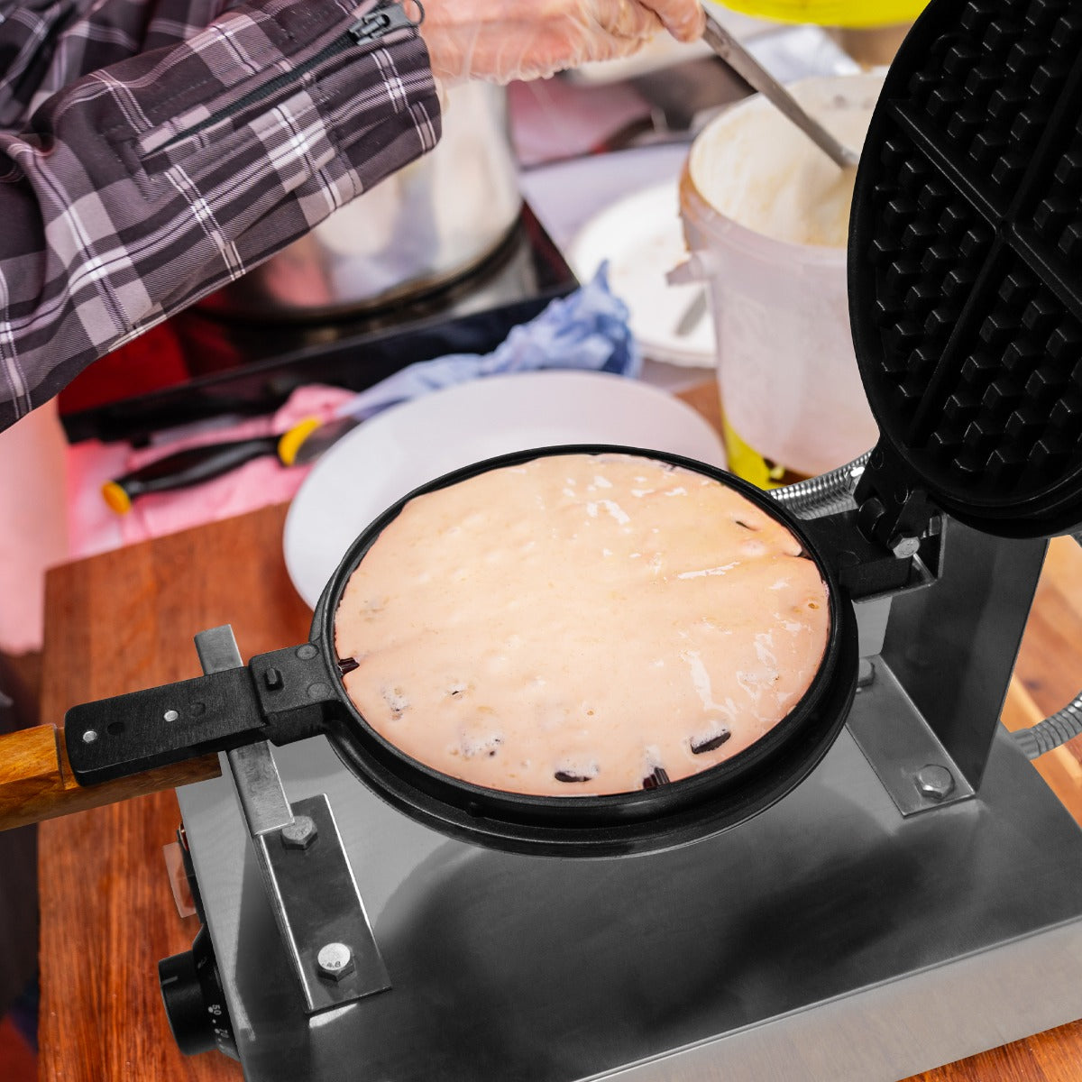 Gaufrier KuKoo en action, pâte à croque-monsieur dorée dans un moule noir, avec une main utilisant une spatule en bois