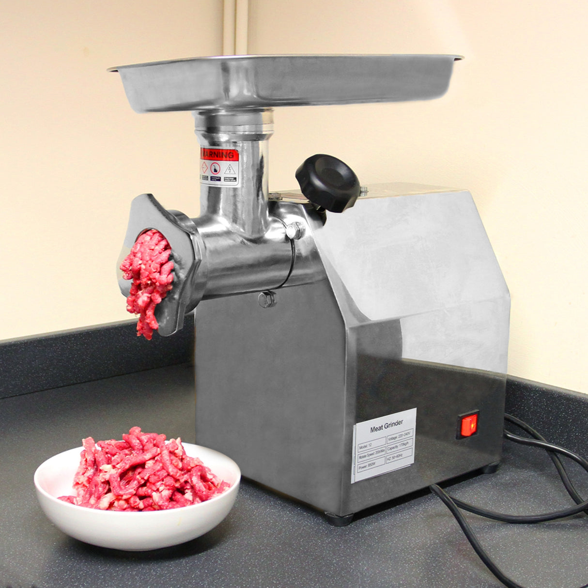 Hachoir à viande commercial en acier inoxydable, avec un bol en plastique rempli de viande hachée rouge, sur un plan de travail noir