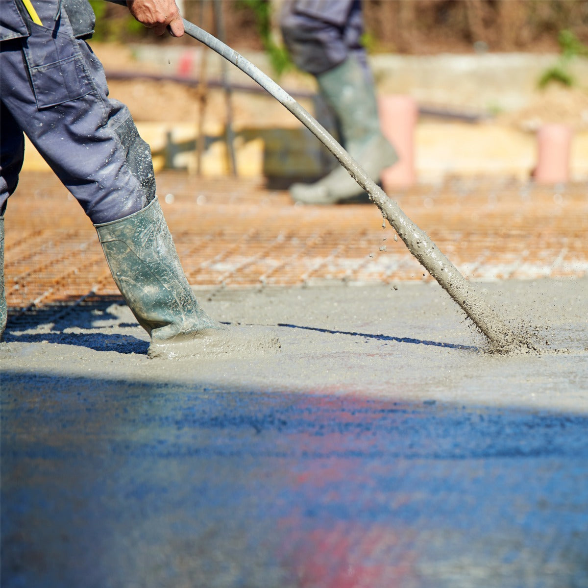 Un ouvrier en bottes en caoutchouc verse du béton frais sur une surface lisse, avec un outil de vibration pour lisser le mélange