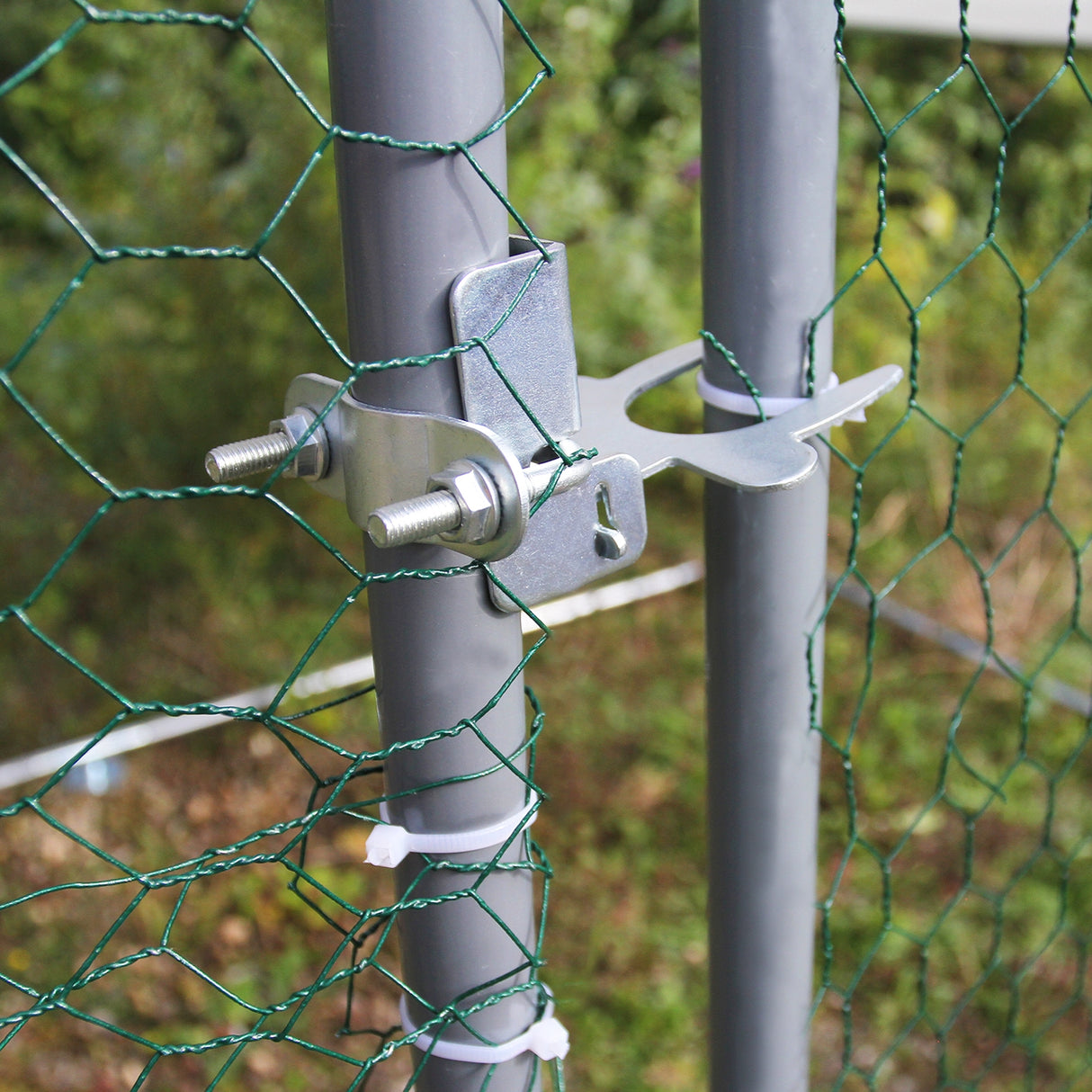 Détail du système de fixation métallique sur un enclos poulailler, avec grillage vert et poteaux en métal gris, en arrière-plan flou