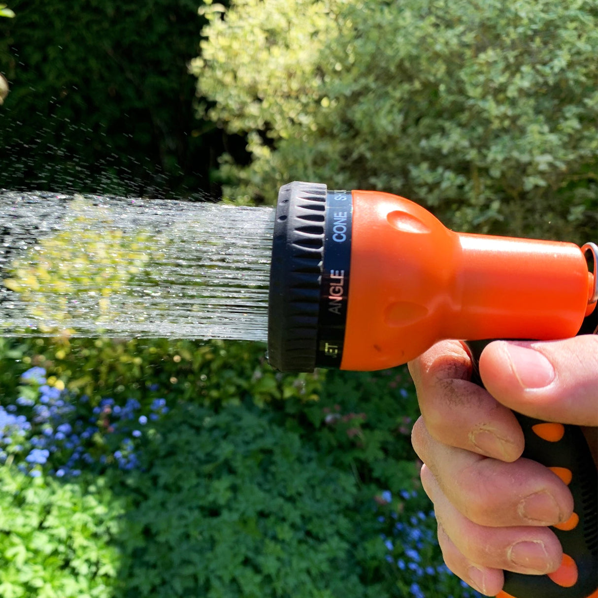 Pistolet d'arrosage orange avec plusieurs réglages, tenu à la main, projetant de l'eau en cône sur un fond de verdure florale