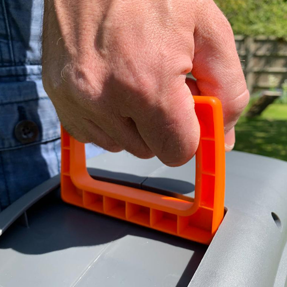 Poignée orange d'un enrouleur de tuyau d'arrosage, main tenant fermement, sur un fond de jardin verdoyant et ensoleillé
