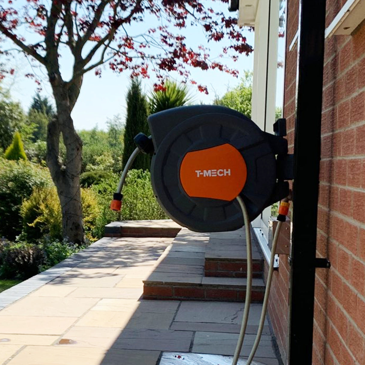 Enrouleur de tuyau d'arrosage noir et orange de 30m fixé à un mur, avec un jardin verdoyant en arrière-plan et un ciel bleu clair