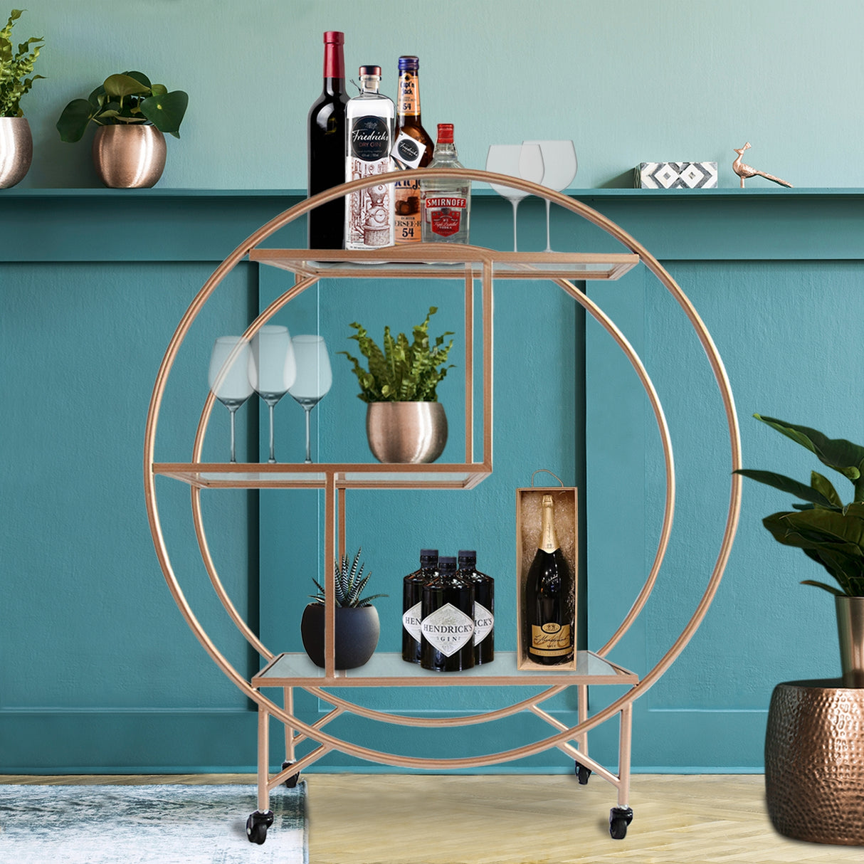 Chariot de bar en or rose, avec étagères rondes, bouteilles de vin, verres à pied et plantes décoratives sur fond turquoise