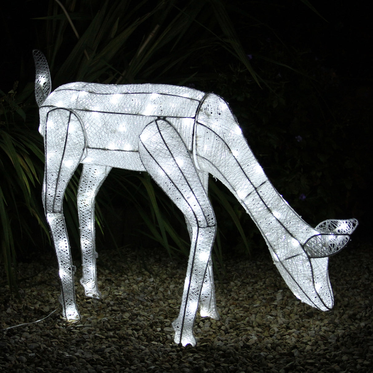 Biche LED en blanc froid, illuminée avec des lumières scintillantes, en position de broutage sur un fond de gravier et de plantes vertes