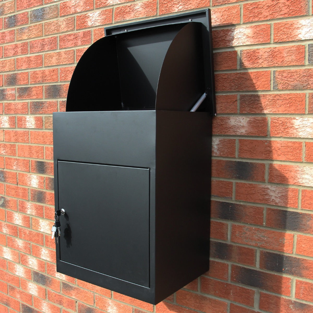 Boîte à colis noire en métal, design moderne avec une ouverture arrondie et une porte verrouillable, fixée sur un mur en briques rouges