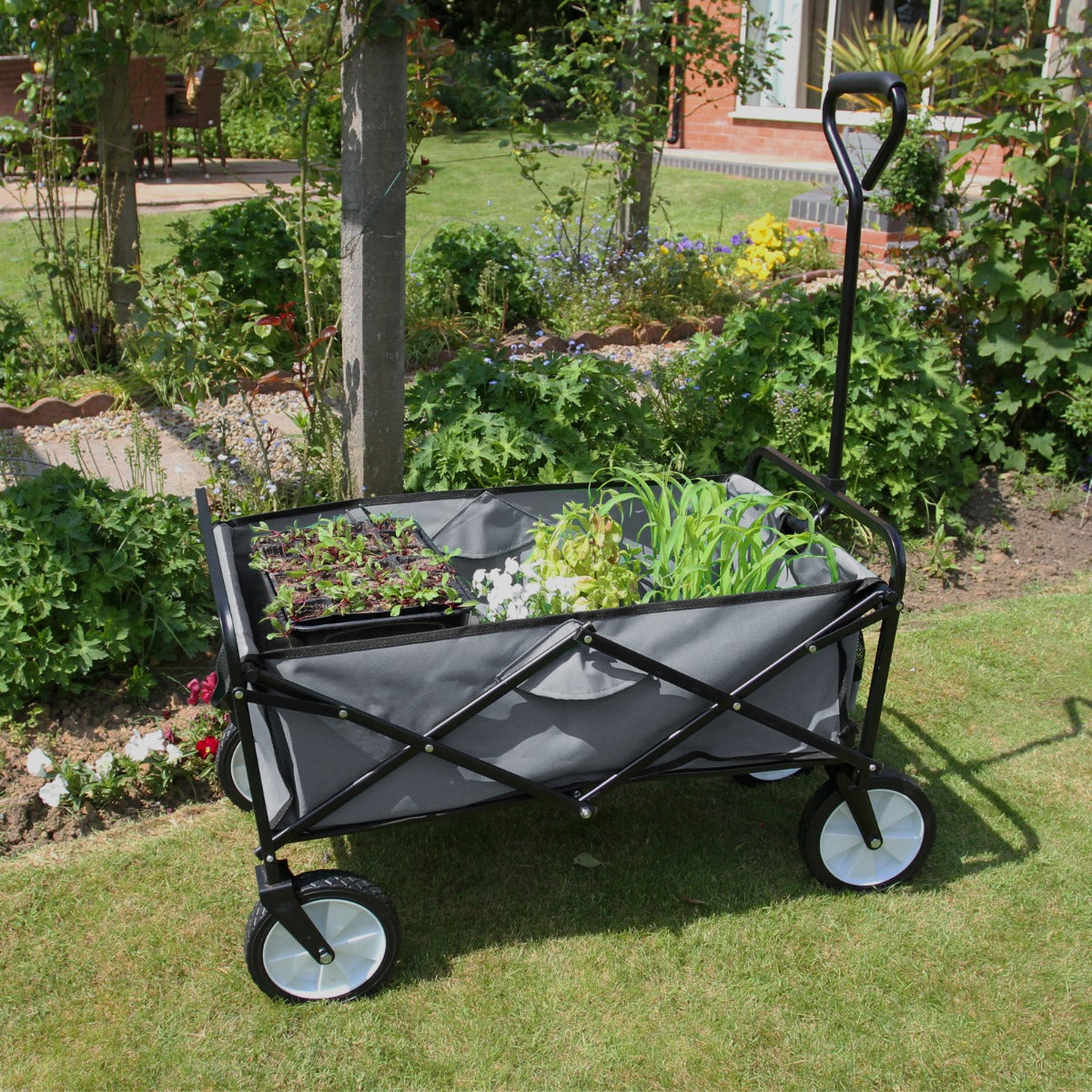 Chariot pliable de jardin gris avec des roues blanches, rempli de plantes vertes et fleuries, sur une pelouse verdoyante