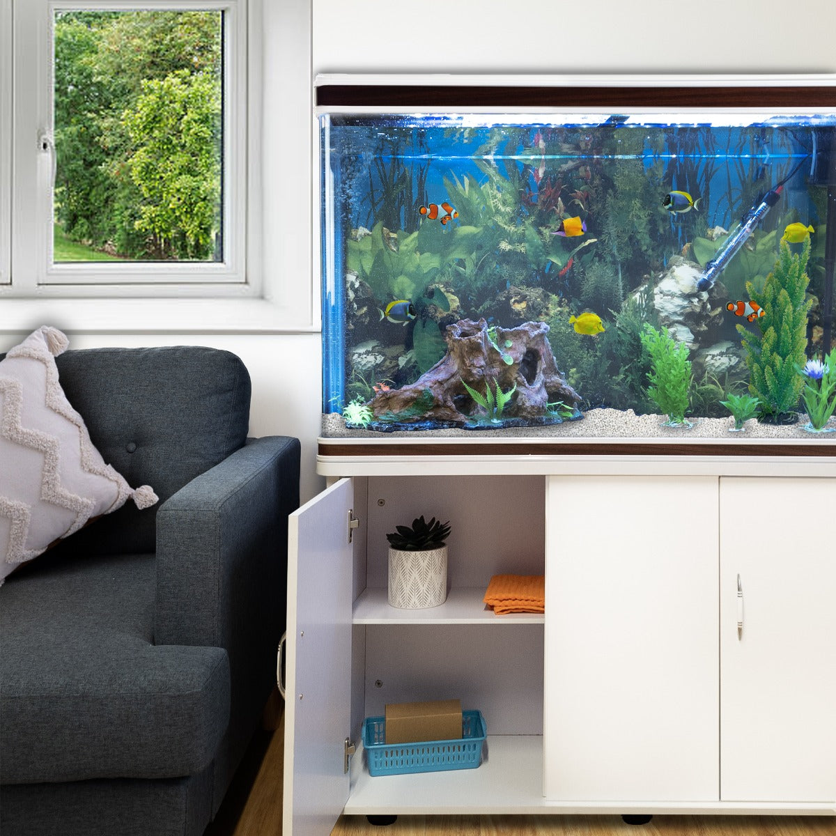 Aquarium blanc avec poissons colorés, meuble de support moderne, gravier blanc, et plante verte, à côté d'un canapé gris et d'une fenêtre
