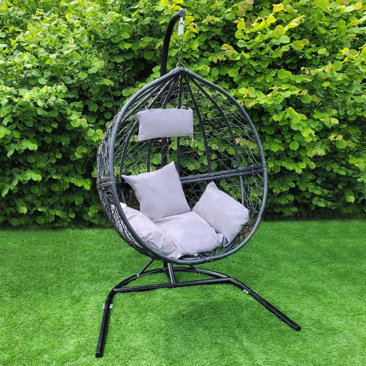 Chaise suspendue en rodin noir, en forme de goutte, avec coussins blancs, sur un support en métal, entourée de verdure luxuriante