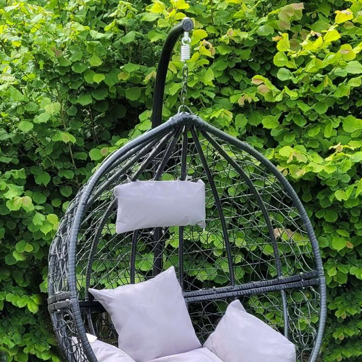 Chaise suspendue en rotin noir avec coussins blancs, suspendue à un support, entourée de verdure luxuriante