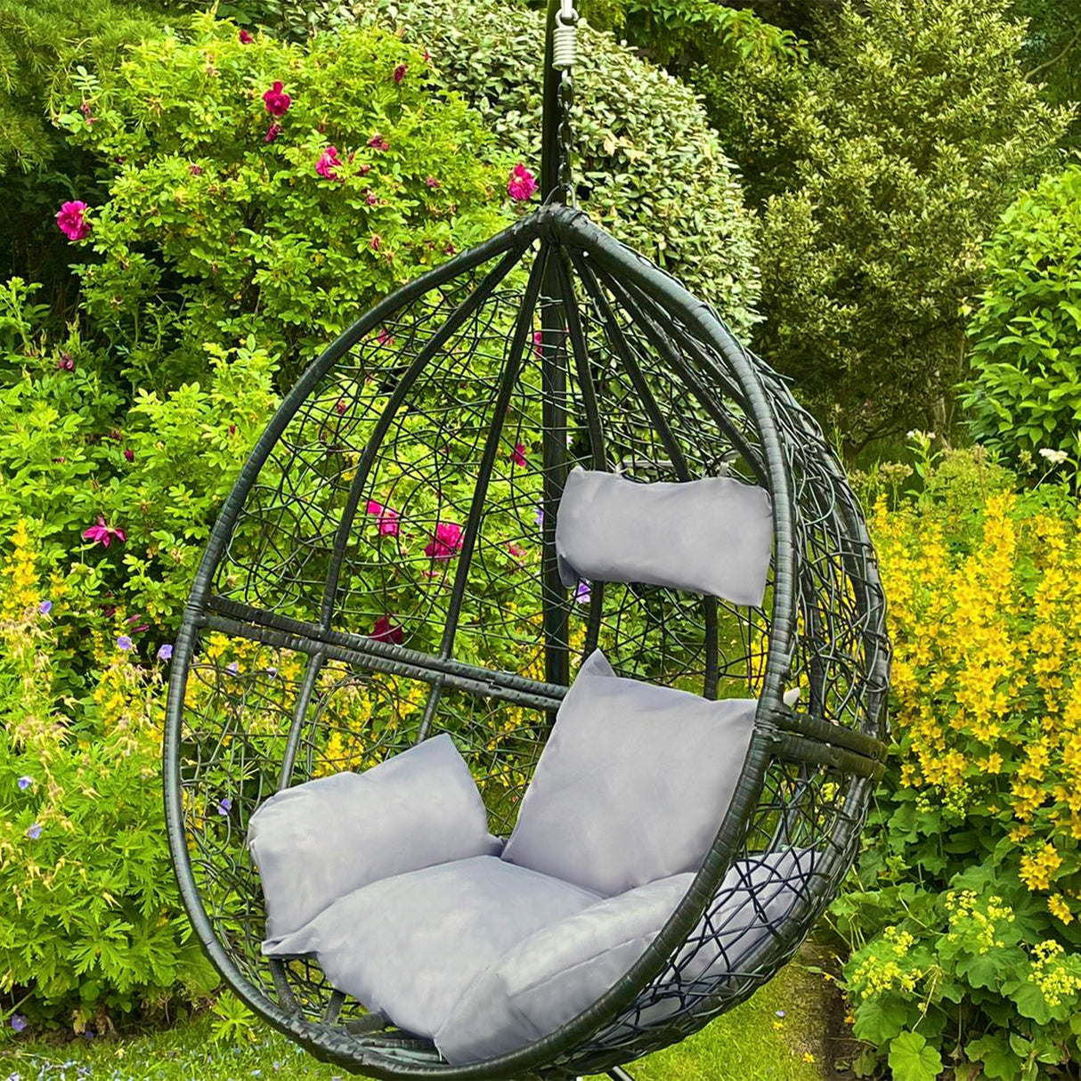 Chaise suspendue en rotin noir avec coussins gris, suspendue dans un jardin verdoyant entouré de fleurs colorées