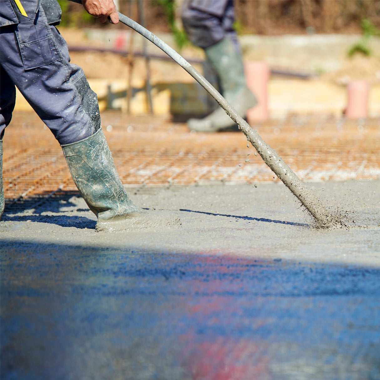 Vibreur à béton électrique en action, avec un ouvrier en bottes en caoutchouc, lissant le béton frais sur une surface grise brillante