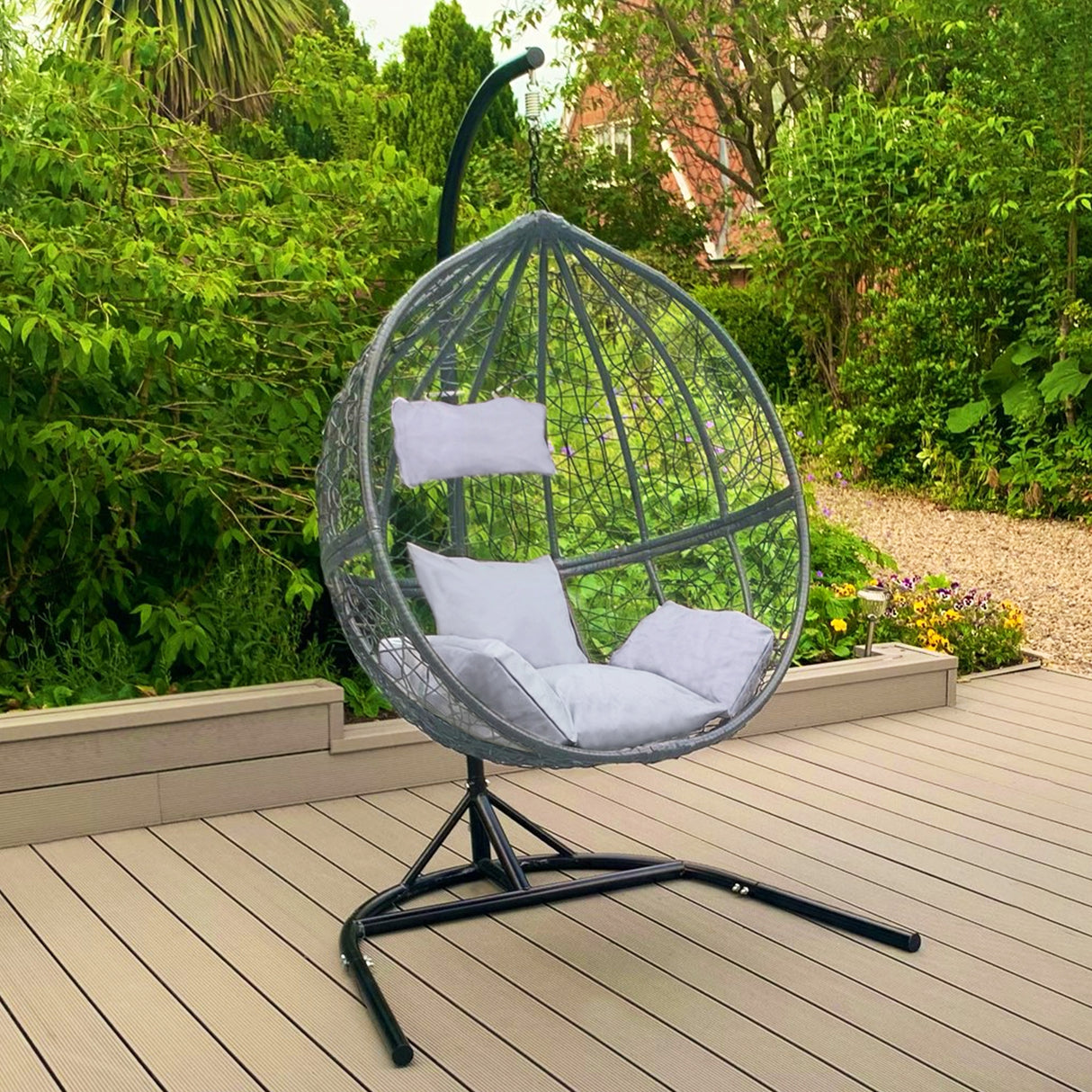Chaise suspendue en rotin gris avec coussins blancs, suspendue sur un support noir, entourée de verdure et d'un sol en bois clair