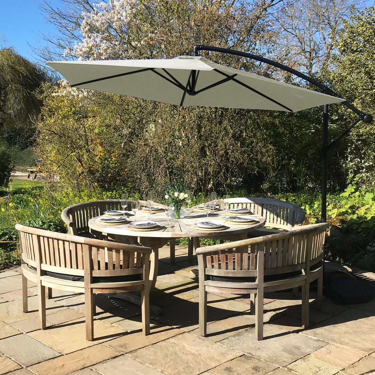 Parasol déporté crème de 3 m sur une table en bois entourée de chaises en bois, dans un jardin verdoyant ensoleillé