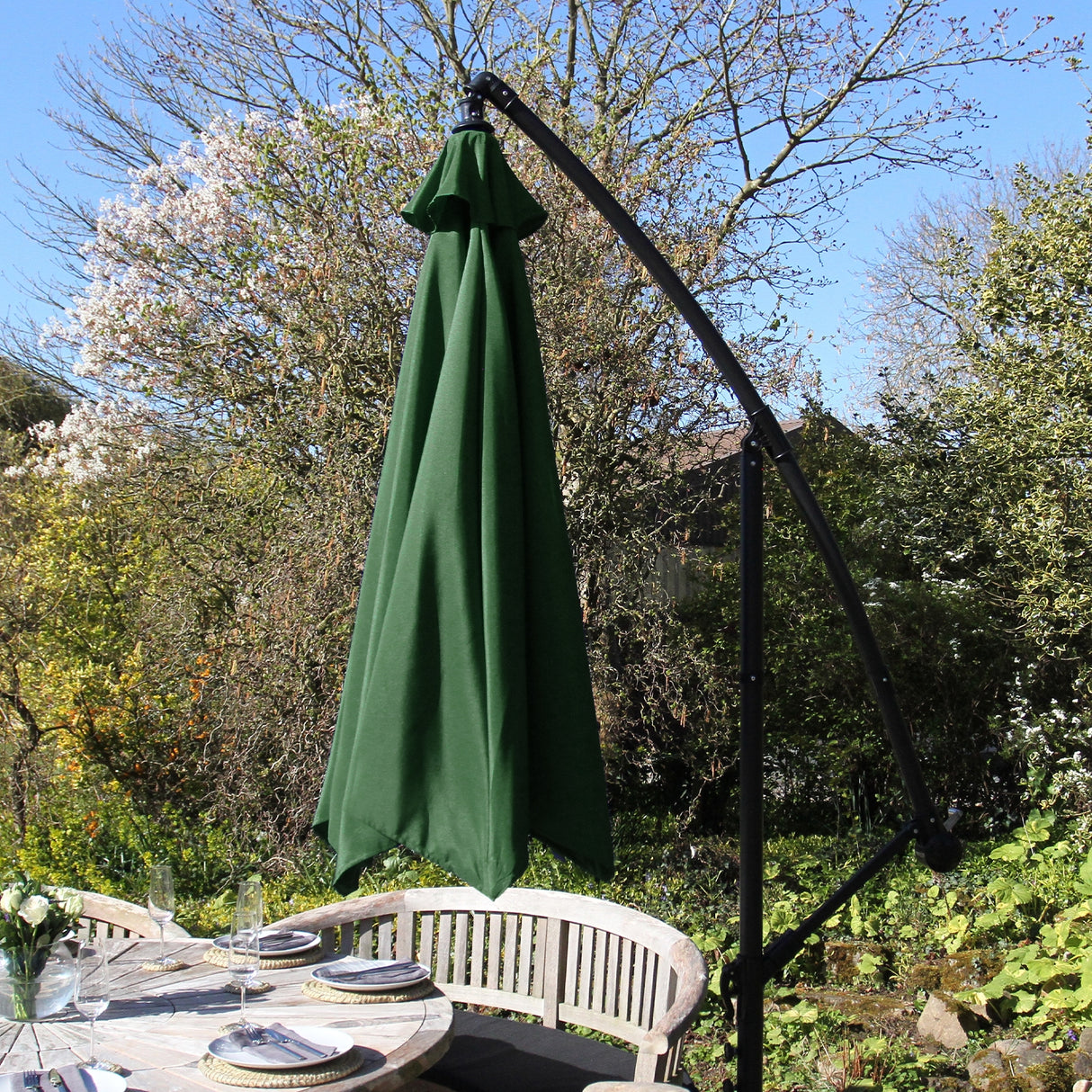 Parasol déporté de 3 m en vert, avec un mât noir élégant, offrant une ombre généreuse sur une table en bois dans un jardin fleuri