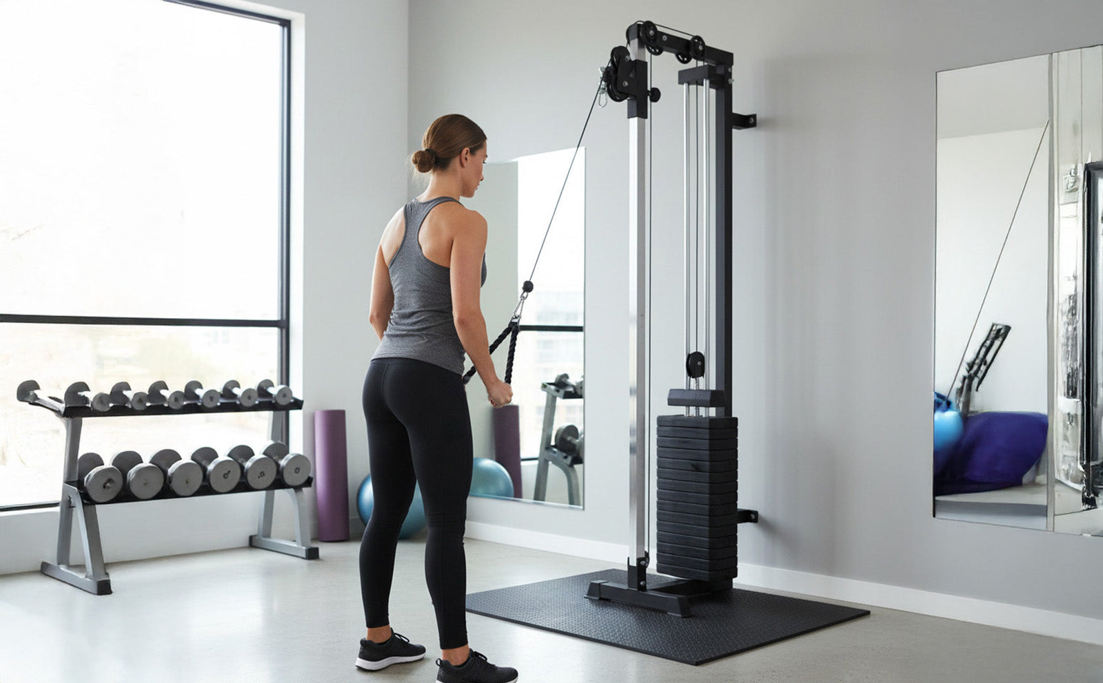 Femme en legging noir et débardeur gris utilisant une station à câbles murales dans un espace de gym moderne, avec miroirs et poids visibles