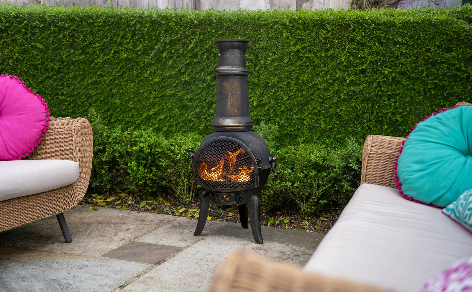 Brasero mexicain en métal noir avec cheminée, flammes vives, entouré de fauteuils en osier et coussins colorés sur un patio verdoyant
