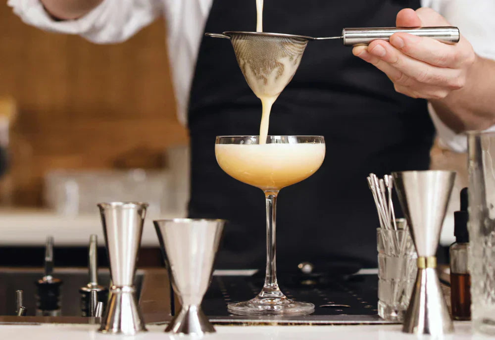 Un barman verse un cocktail crémeux dans une coupe élégante, entouré d'ustensiles de bar en acier inoxydable brillants