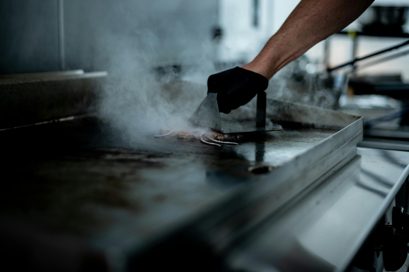 Main d'un cuisinier utilisant une spatule sur une plaque électrique, avec de la vapeur et des aliments cuits sur une surface en métal brillante