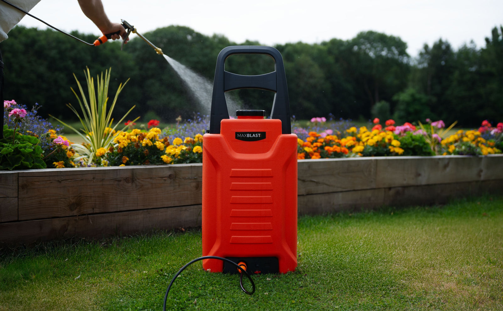 Chariot d'arrosage rouge vif, avec un tuyau noir, devant un jardin coloré de fleurs, idéal pour les jardiniers paresseux