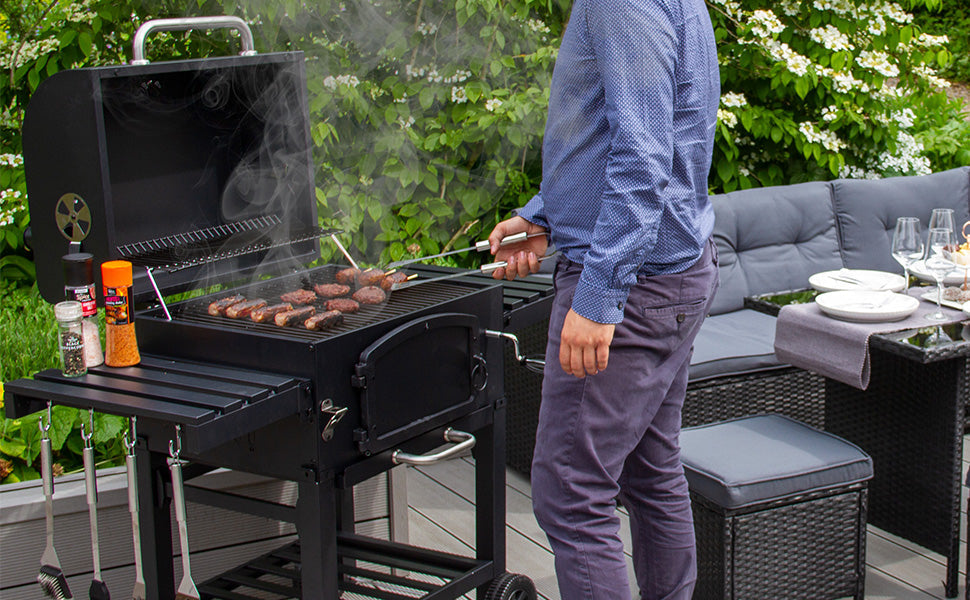 Homme en chemise bleue cuisinant des saucisses sur un barbecue noir, fumée s'échappant, avec un jardin verdoyant en arrière-plan