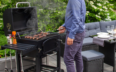 Homme en chemise bleue cuisinant des saucisses sur un barbecue noir, fumée s'échappant, avec un jardin verdoyant en arrière-plan