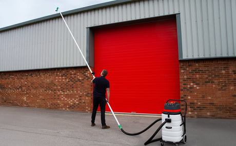 Homme utilisant un aspirateur industriel avec un long tuyau pour nettoyer des gouttières devant un grand garage rouge en métal