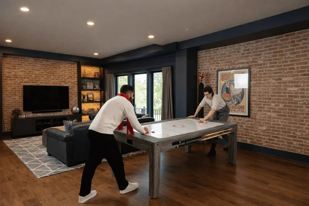 Deux personnes jouent à un air hockey sur une table de jeux 7-en-1, entourées d'un salon moderne avec des murs en briques et un éclairage chaleureux