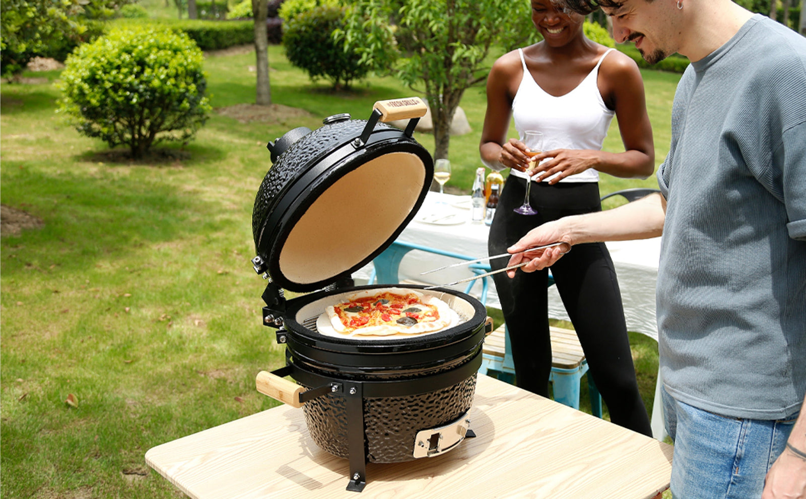 Gril en céramique Kamado noir, avec pizza garnie, utilisé par un homme et une femme dans un jardin verdoyant et ensoleillé